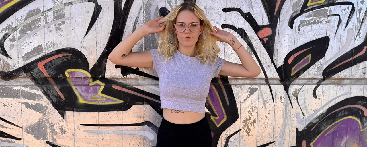 BeiKatha mit rosa Brille im grauen Crop-Top vor einer Graffiti-Wand