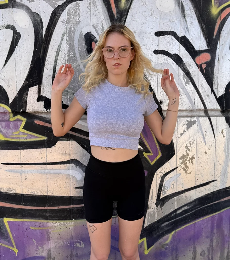 BeiKatha mit rosa Brille im grauen Crop-Top und Radlerhose vor einer Graffiti-Wand